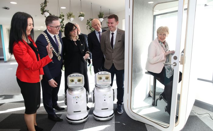 11Minister for Rural and Community Development, Heather Humphreys TD, sits in a white booth with a glass door on a chair with a laptop. Outside of the booth 2 small robots with cat faces on their screen stand with three men and two women looking in at Minister Humphreys at the launch of major funding to support remote working.
