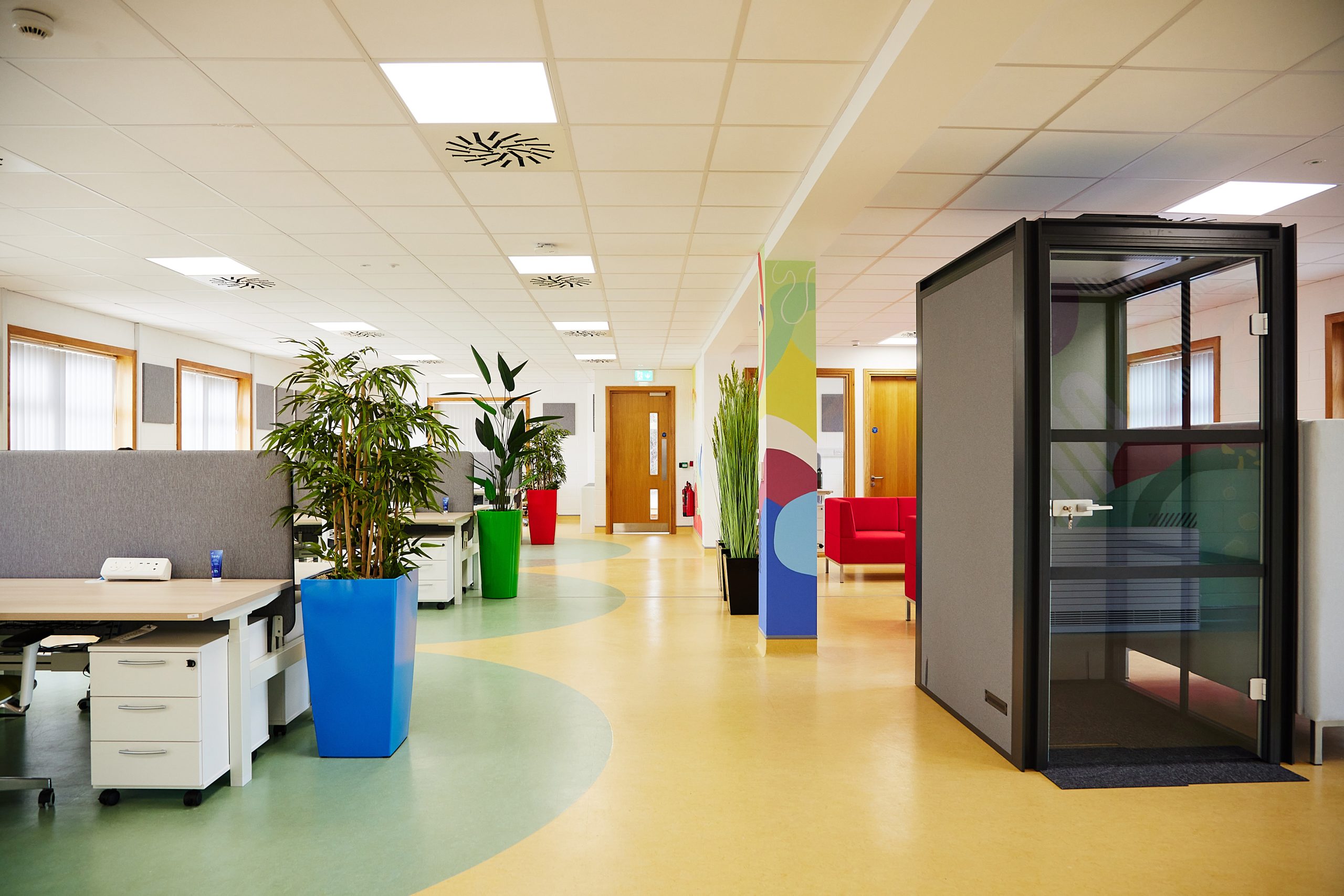 11A bright spacious room, with a bank of desks to the left and a black phone booth and red couches to the left. Plants line the centre walkway along a green and yellow vinyl floor.